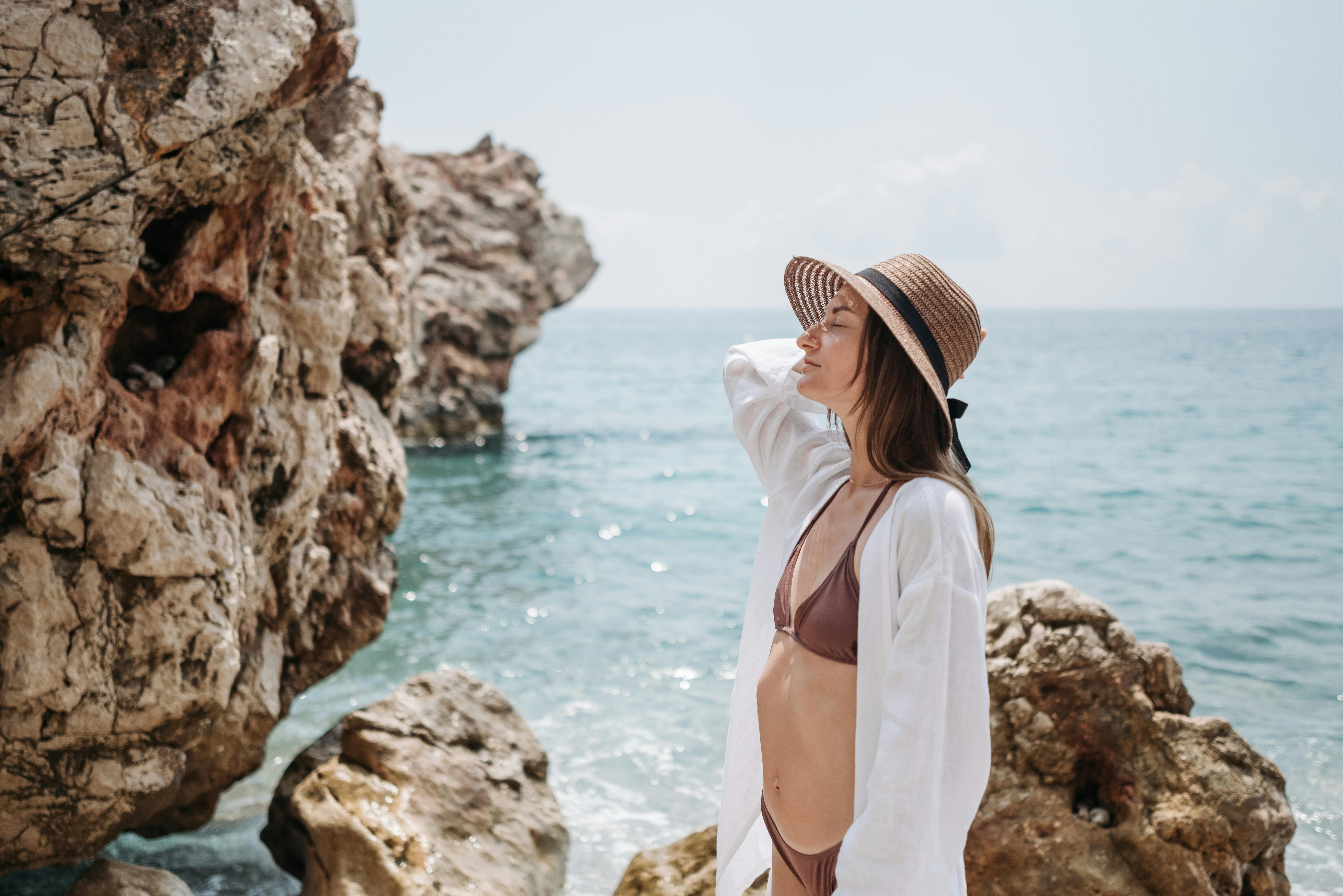Free Woman in swimwear and sun hat enjoying the rocky seaside on a sunny day. Stock Photo