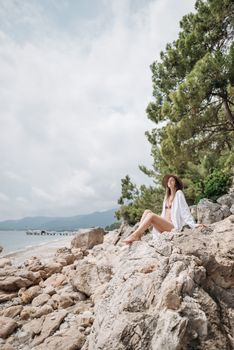 Woman in swimsuit and sun hat sitting on rocky shore enjoying summer at the beach.