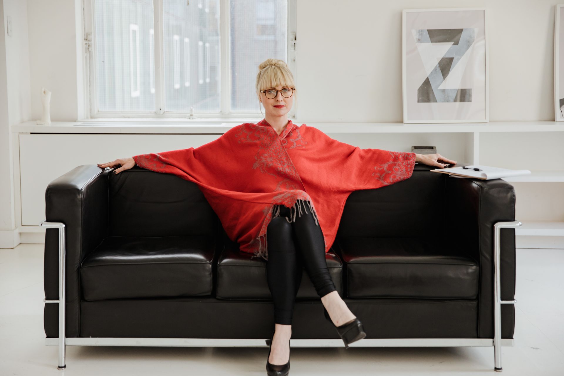 Stylish woman in a red poncho sitting on a modern black sofa in a minimalist indoor setting.
