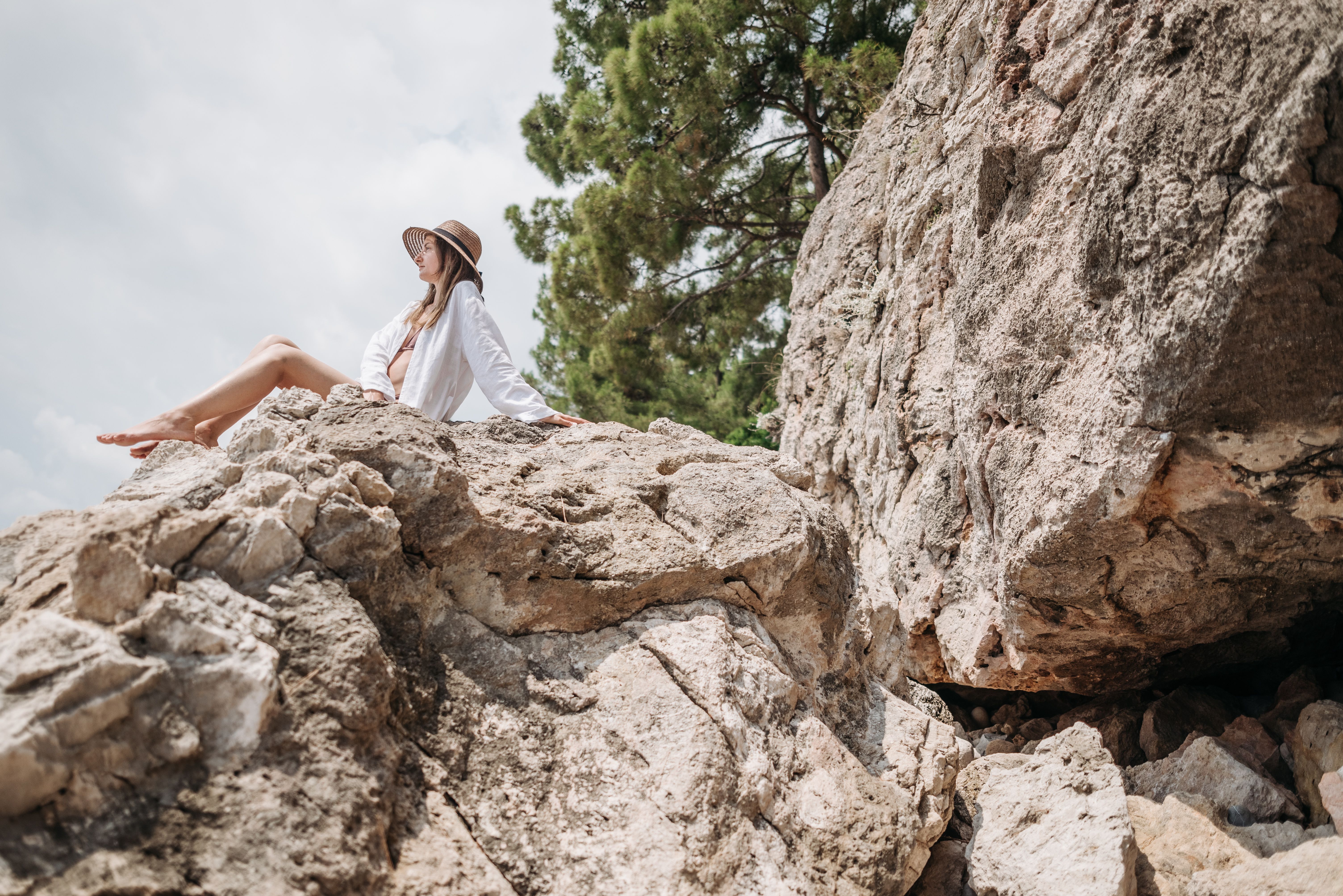A woman in white long sleeves and hat relaxes on a rock with a scenic view.