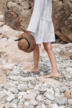 Barefoot woman in a white robe holding a straw hat, walking on rocky beach.