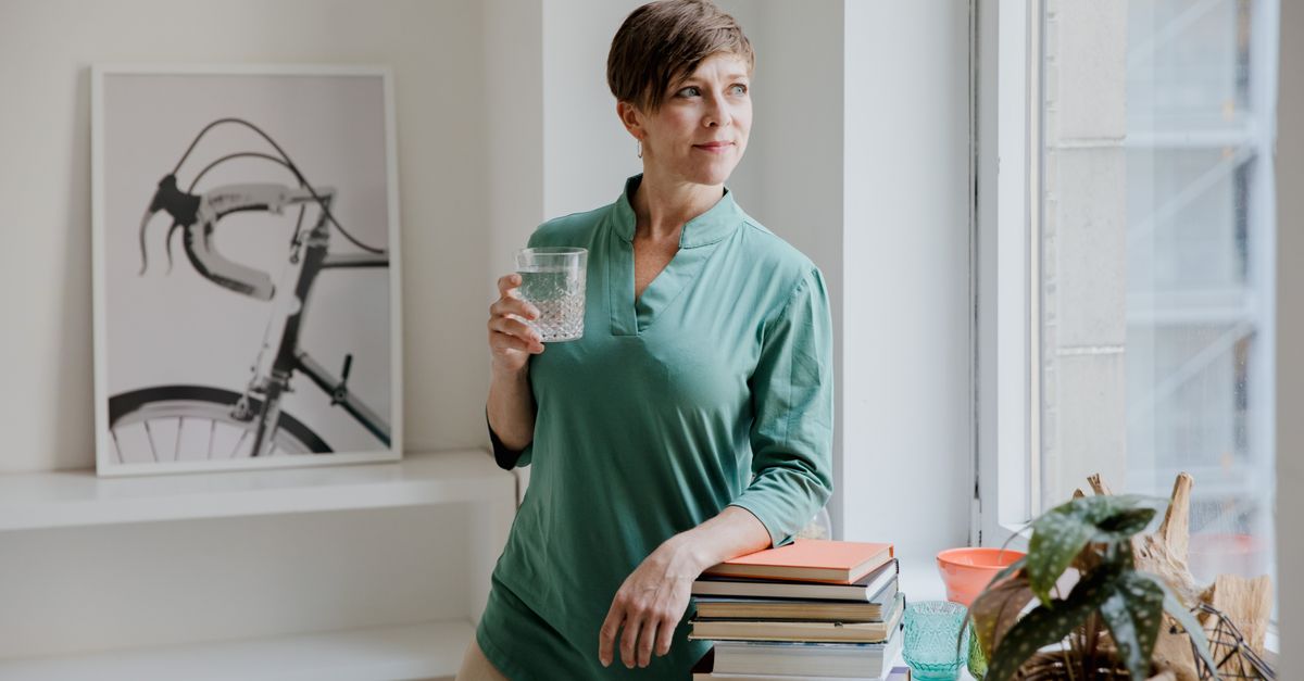 person standing in a bright, airy room with open books and a laptop on a wooden table, looking thoughtful