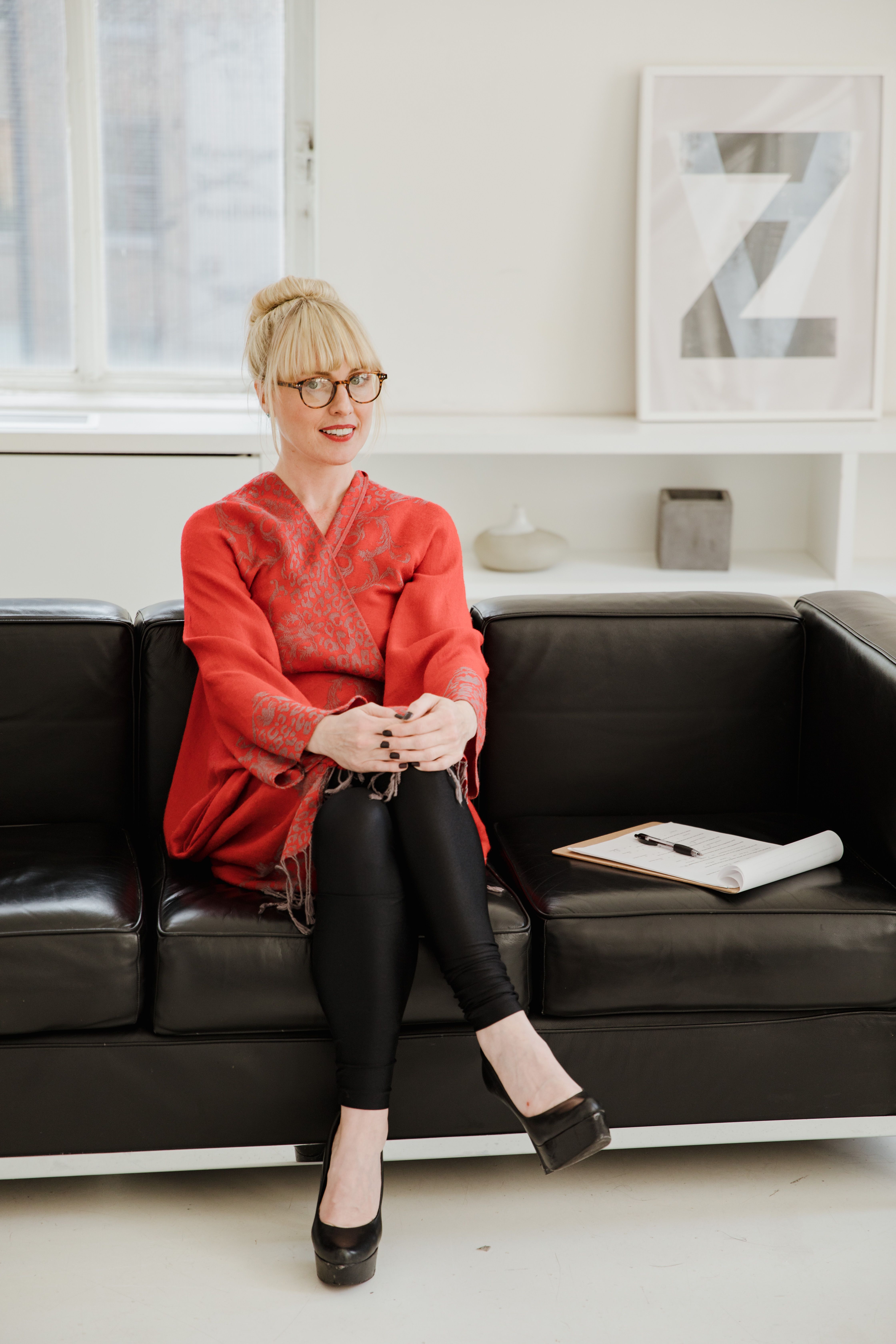 Portrait of a smiling woman in red attire sitting on a black sofa indoors, exuding elegance and style.