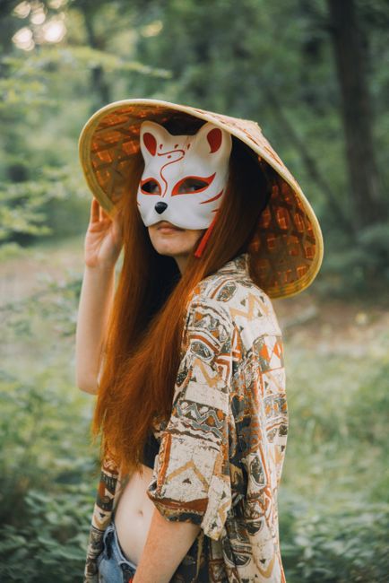 Woman with fox mask and straw hat in serene forest setting, embodying mystique.
