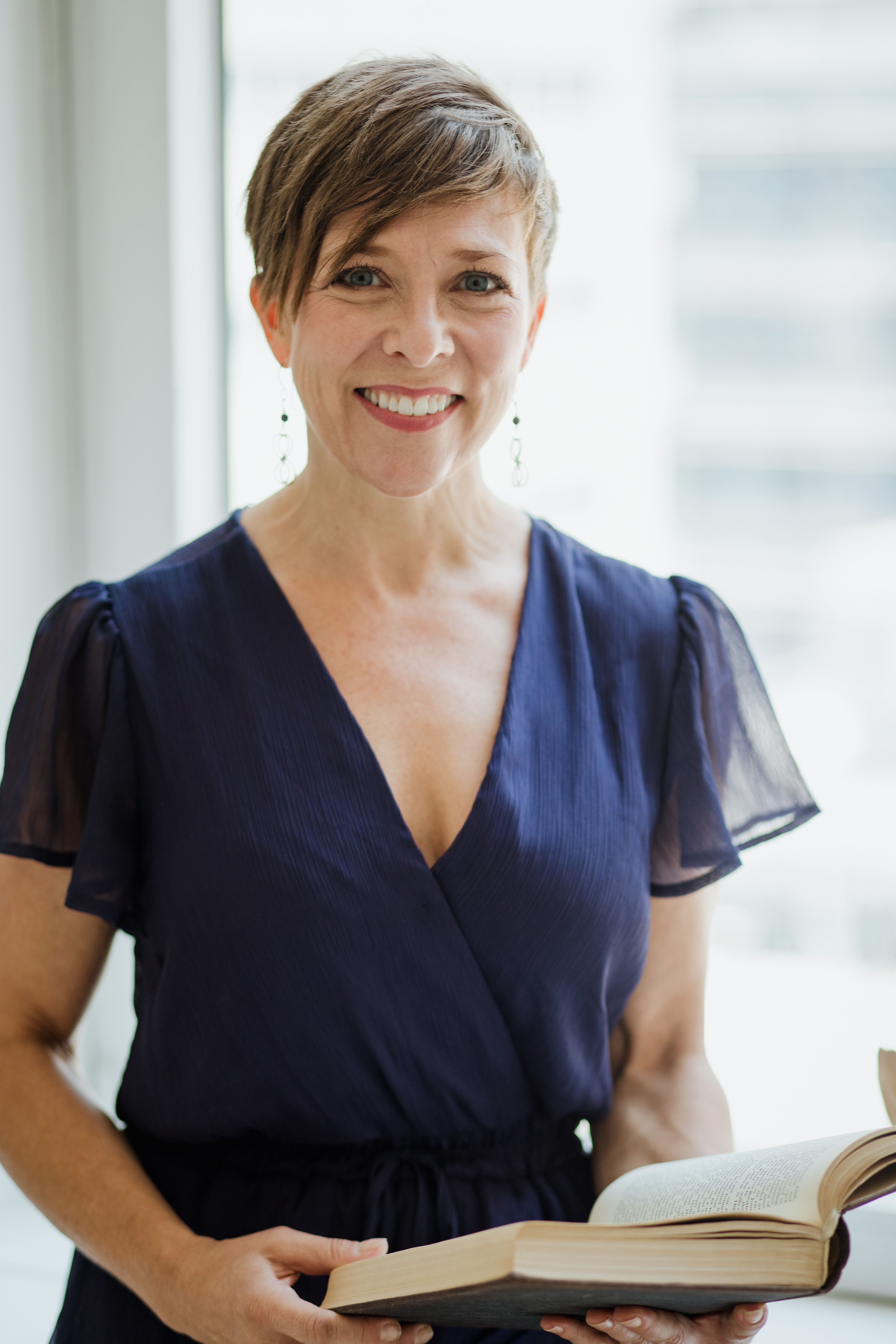 Charming woman with short hair in a blue dress smiling while holding an open book.
