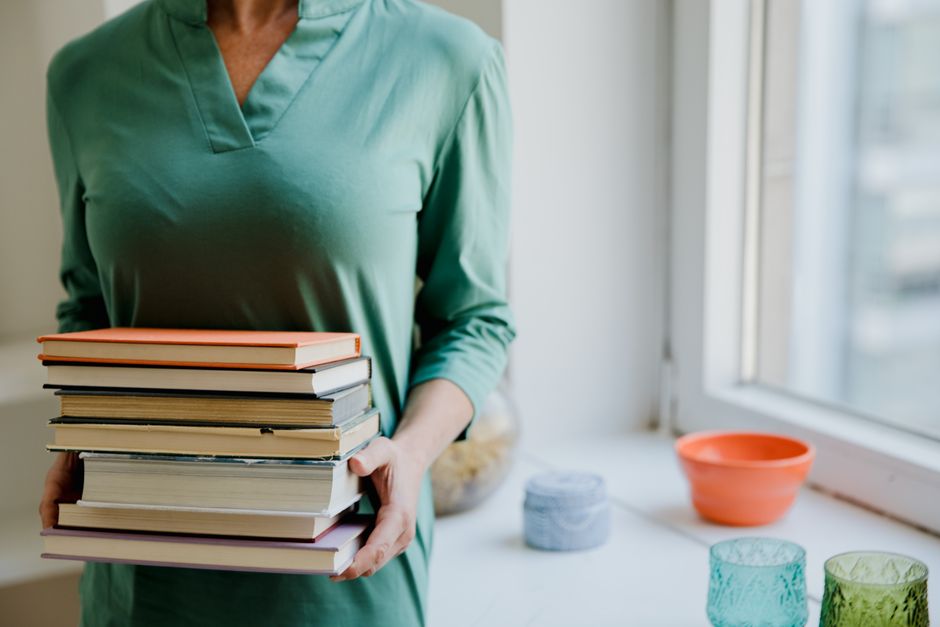A person in a green dress holds a stack of books indoors by a window.