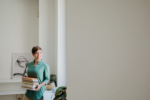 Free A cheerful woman with short hair stands indoors holding books, looking thoughtfully out the window. Stock Photo