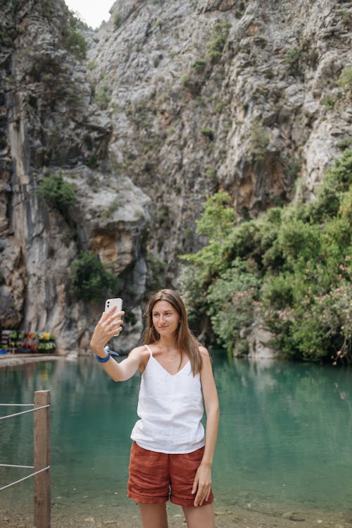 A Woman in a Spaghetti Strap White Top Taking a Selfie beside a River