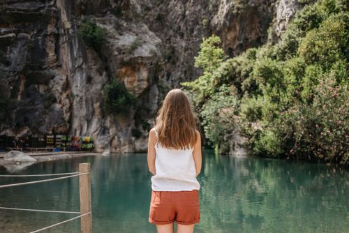 Free A woman stands facing a tranquil lake surrounded by rocky cliffs and lush greenery. Stock Photo