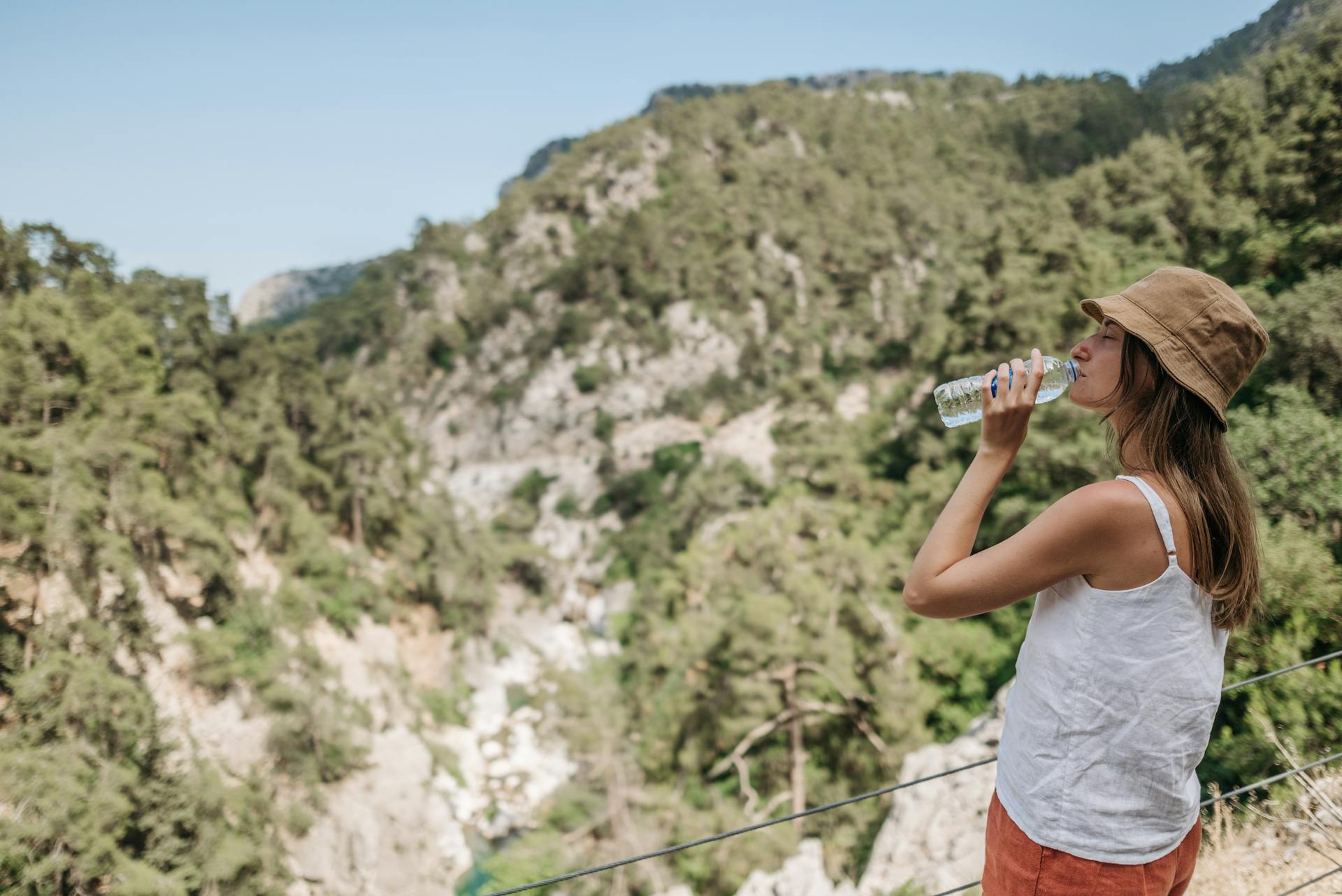 Woman in a bucket hat drinking water while hiking in a picturesque canyon.