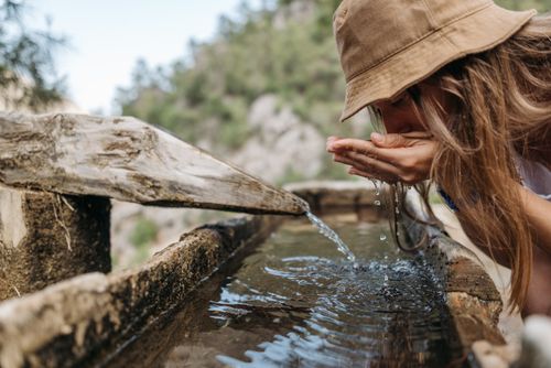 Free A young woman in a hat drinks from a rustic outdoor fountain in nature. Stock Photo