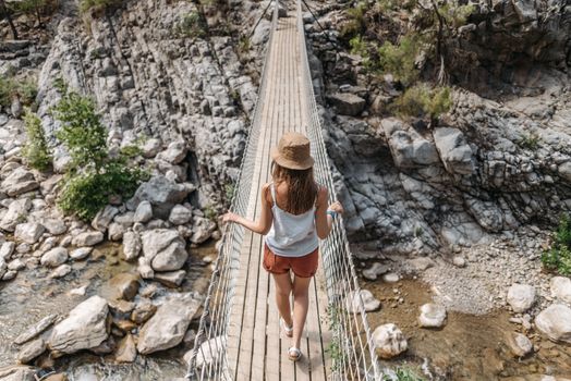 A woman in casual attire crosses a hanging bridge over rocks in a scenic outdoor setting.