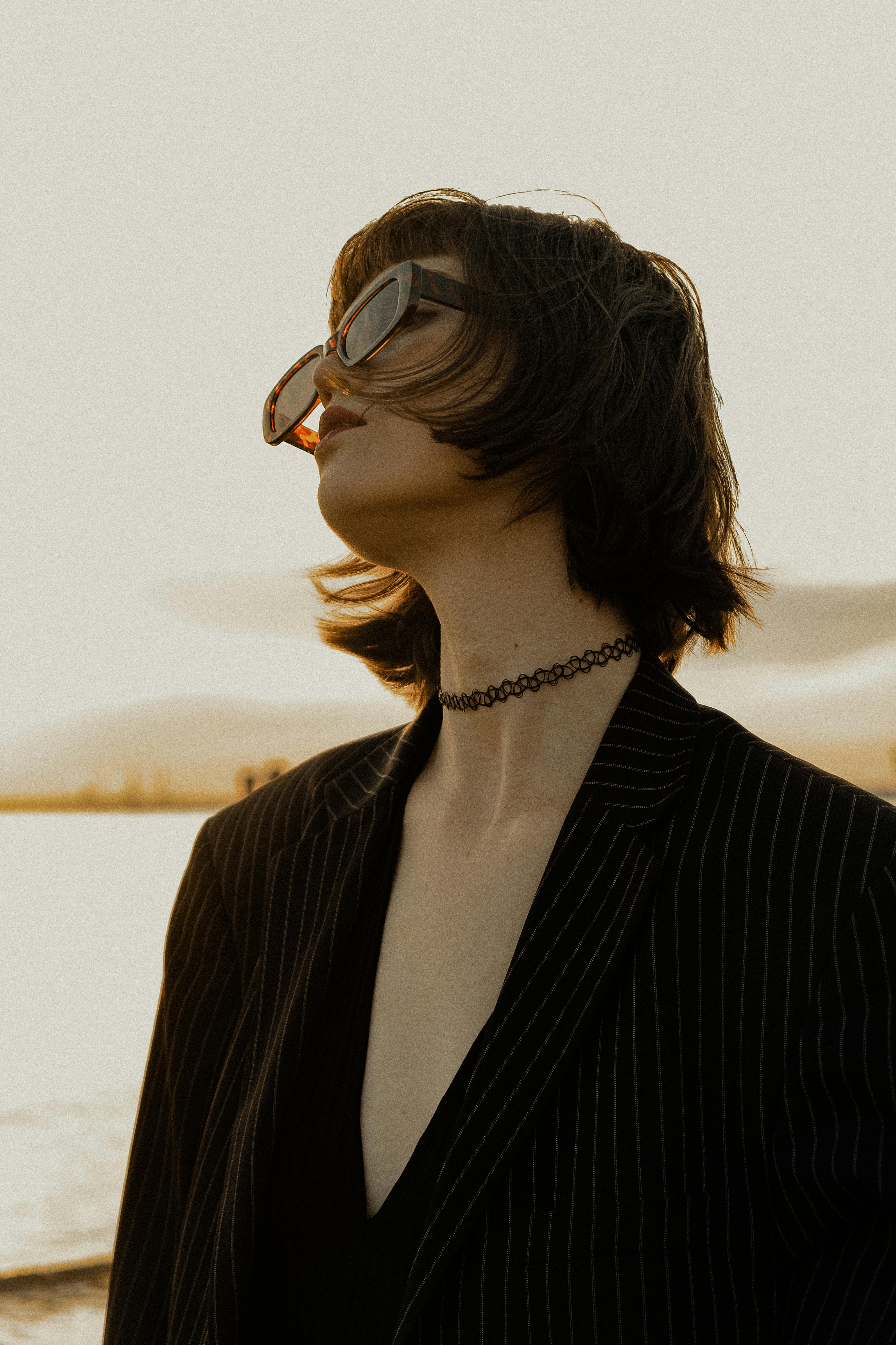 Stylish woman in sunglasses and pinstripe blazer posing by the water at sunset.
