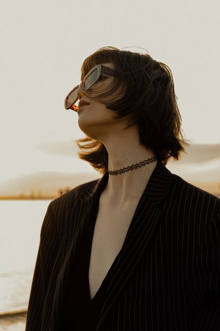 Stylish woman in sunglasses and pinstripe blazer posing by the water at sunset.
