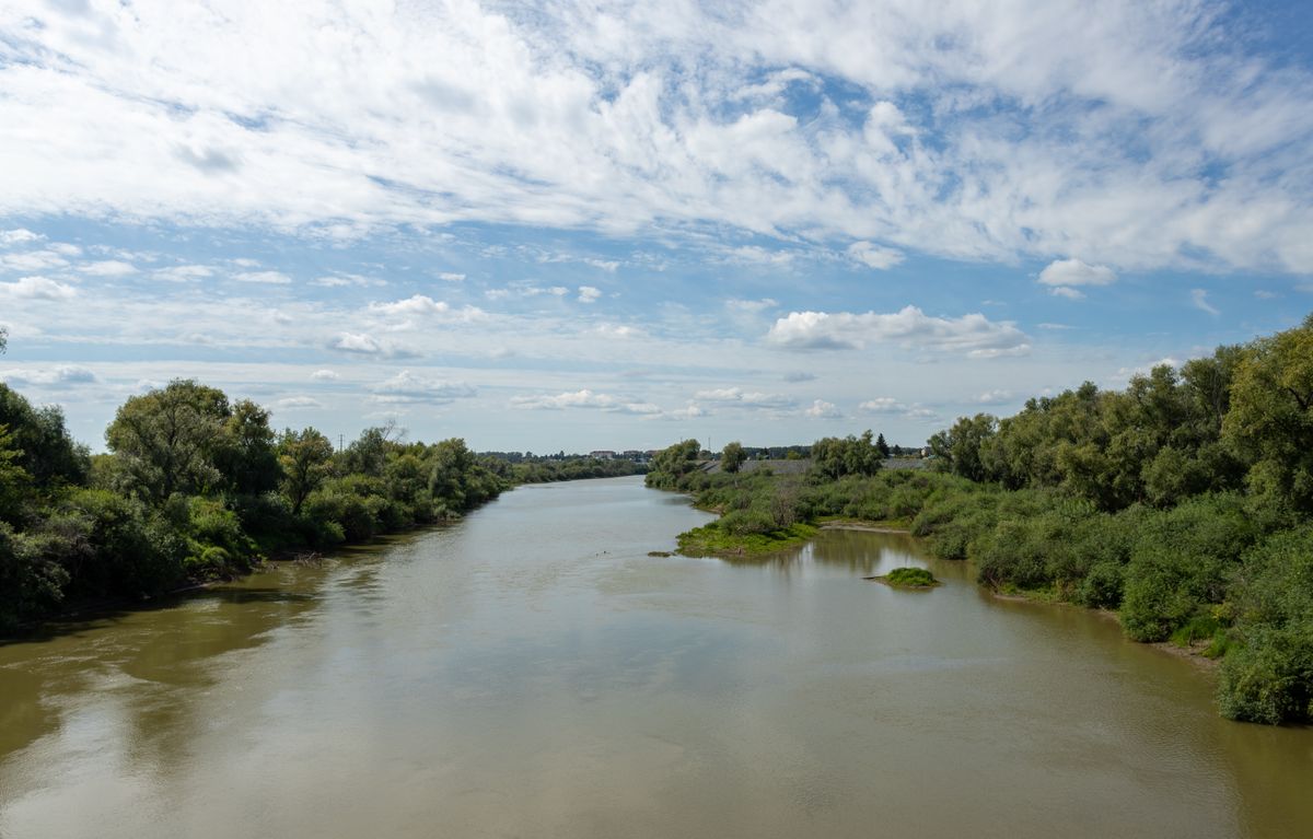 Broad, slow, muddy river runs between low willow-lined banks under a big prairie sky