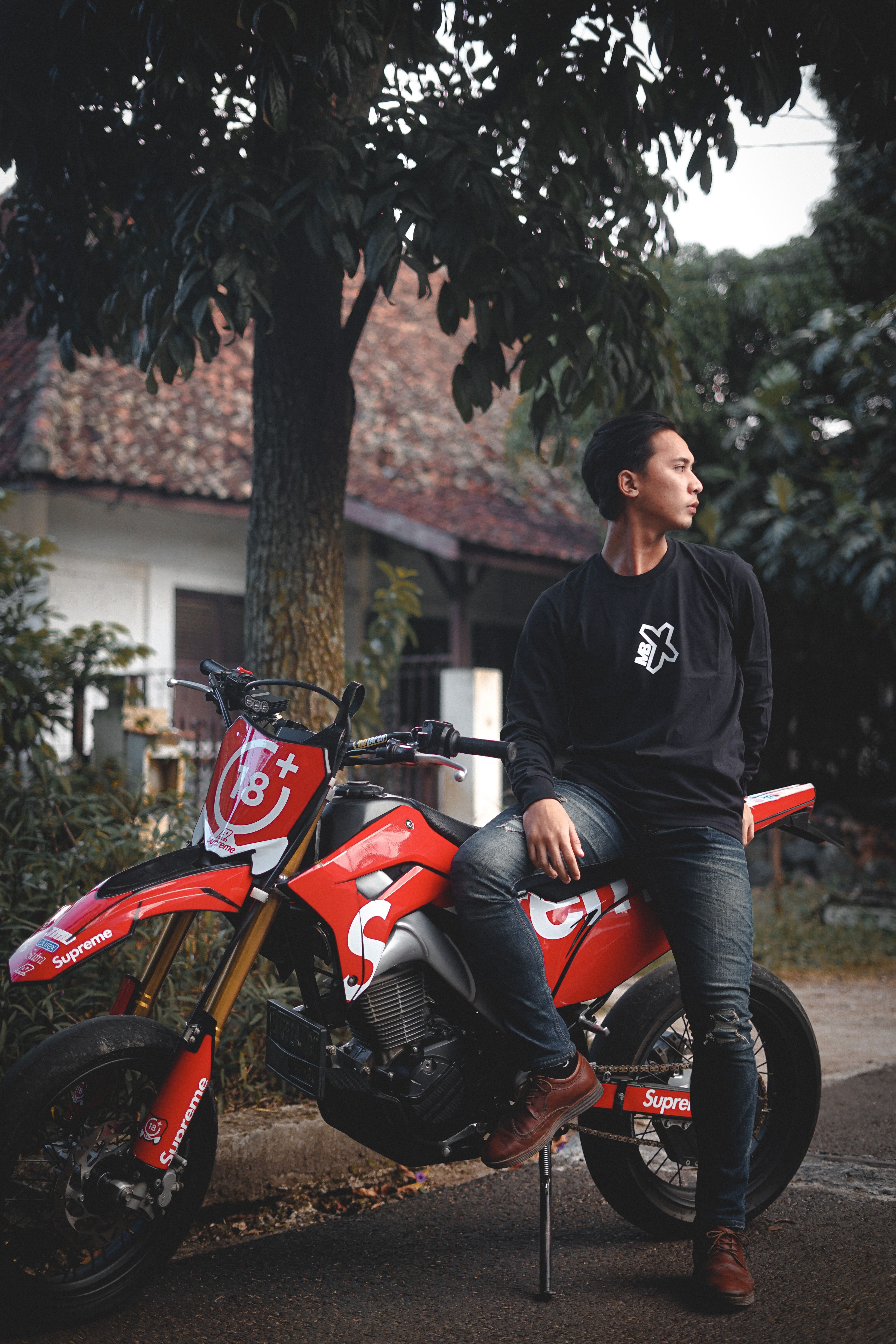 A young man in a black sweater poses confidently on a red motorcycle, set in a leafy outdoor area.