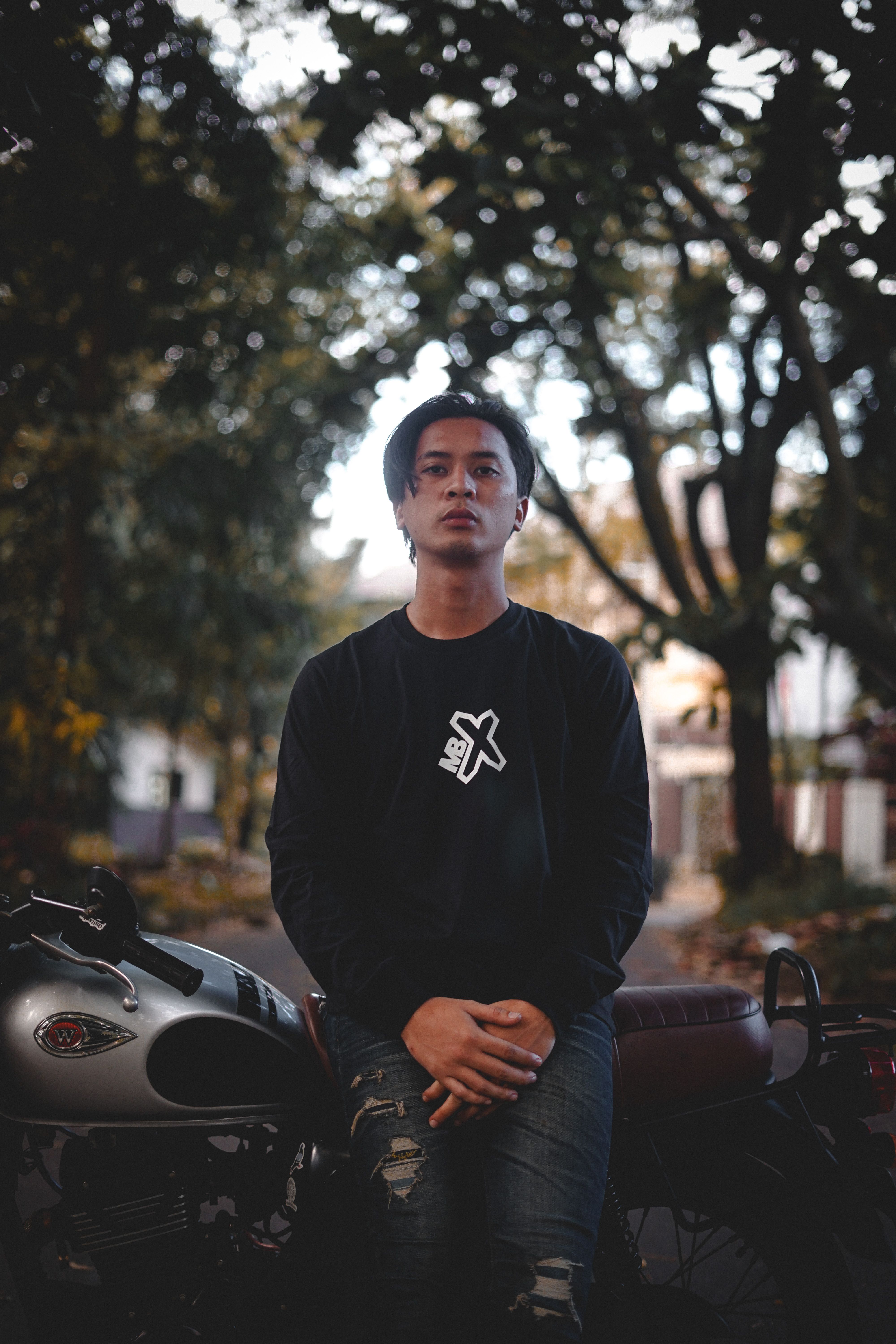 A young man sits on a classic motorcycle surrounded by trees in an outdoor setting.