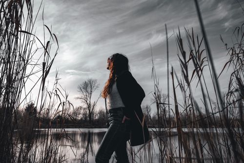 Free Side profile of a woman in nature, wearing glasses, with wild grass and a lake backdrop under a cloudy sky. Stock Photo