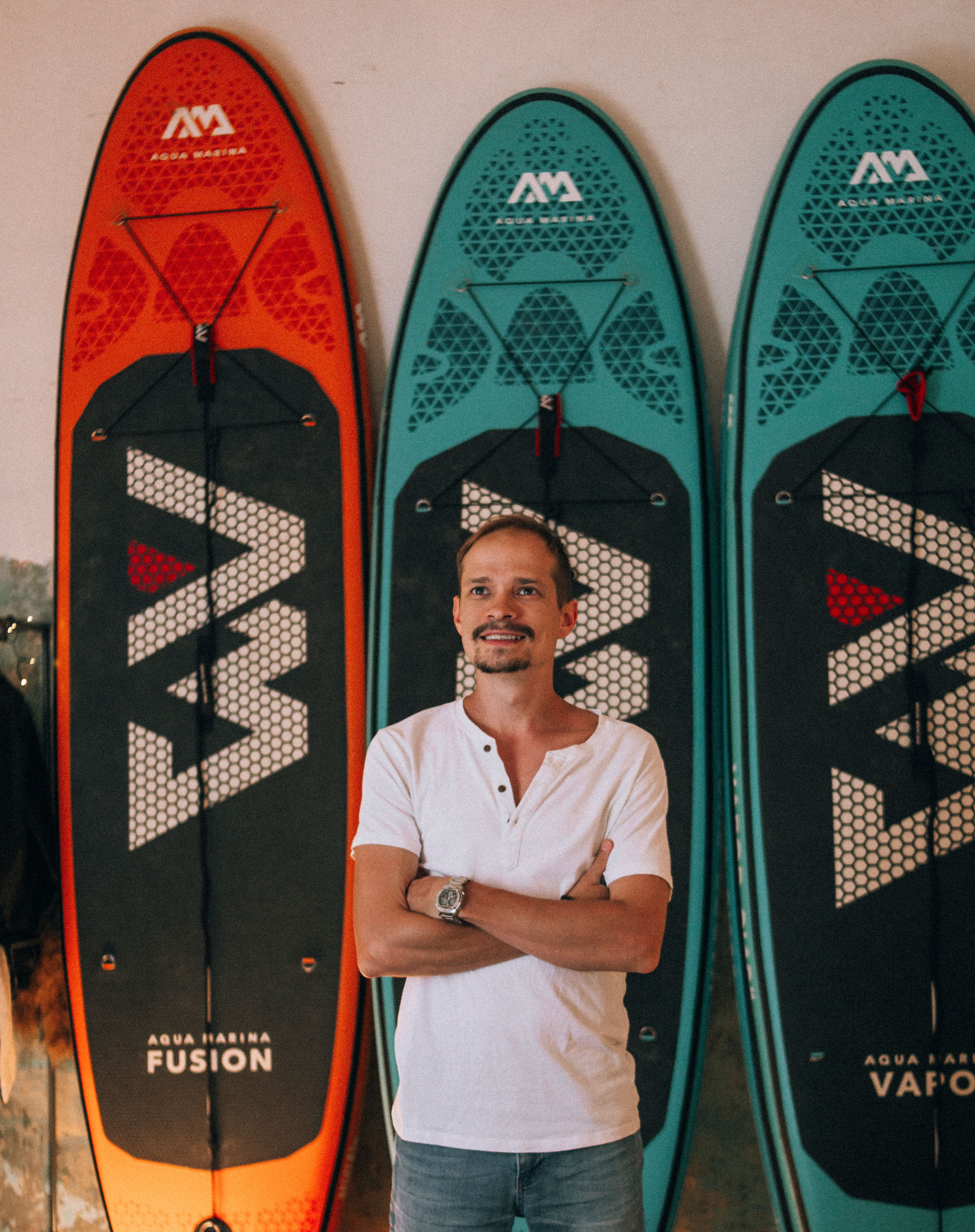Smiling man in white shirt posing indoors with vibrant surfboards behind him.