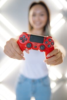 A person holds a red game controller towards the camera with a bright blurred background.