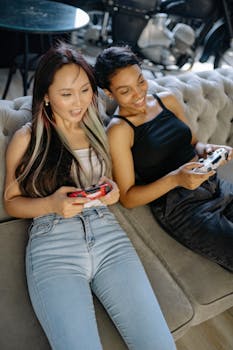 Two women playing video games and enjoying their time indoors on a sofa.