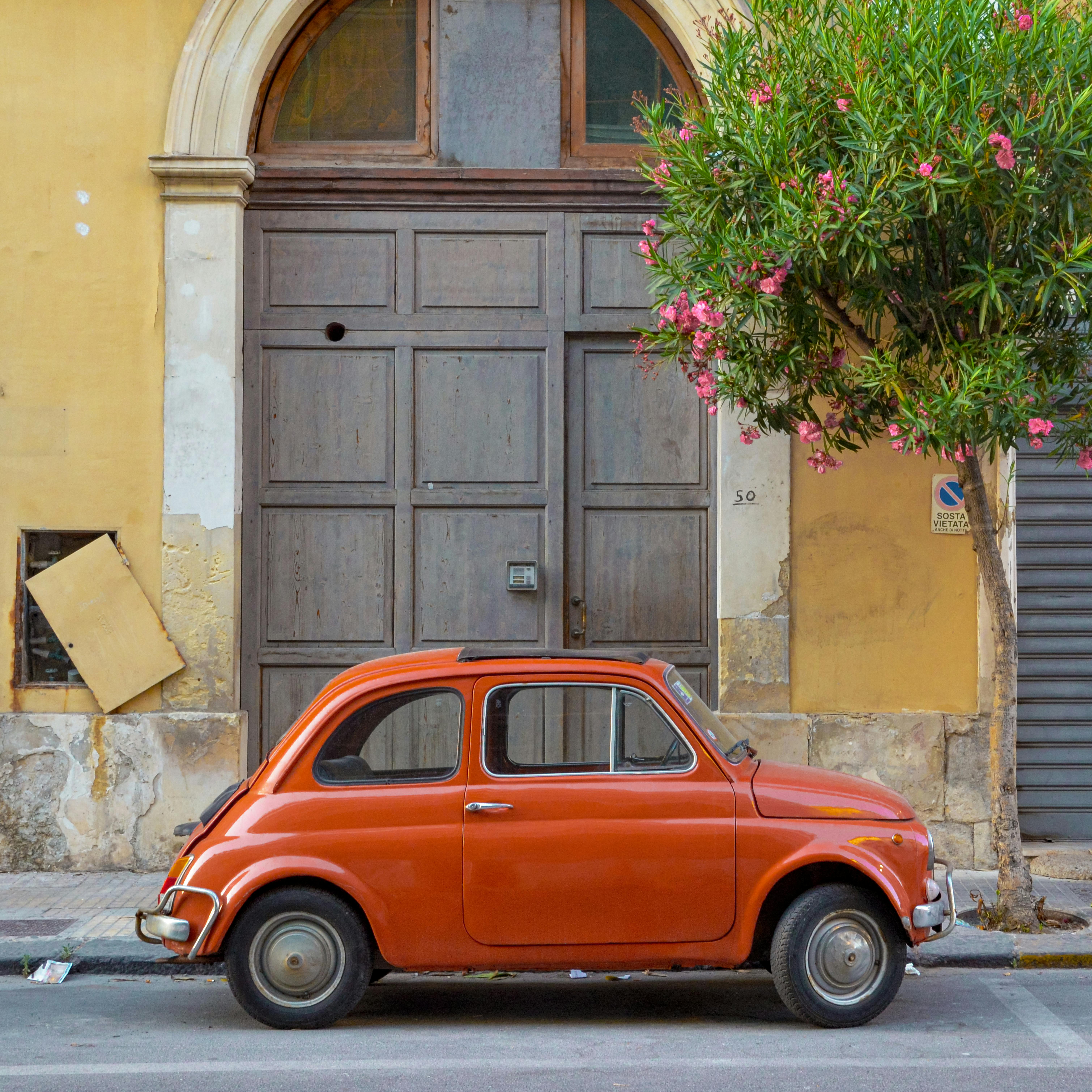 Classic orange Fiat parked near a wooden door in Sicily, Italy. Charming street scene.