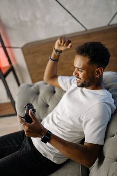 A man happily plays video games on a stylish sofa in a modern living room.