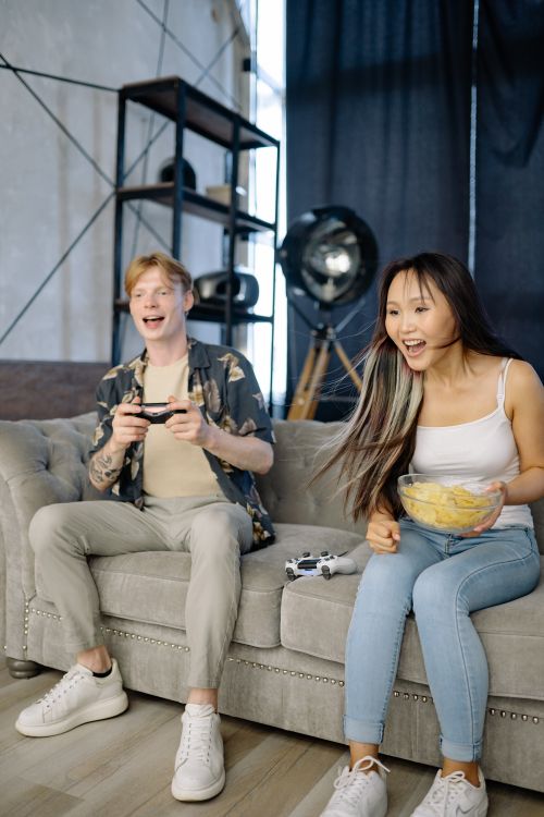 Free Asian woman and Caucasian man laughing while gaming on the couch, enjoying quality indoor time. Stock Photo