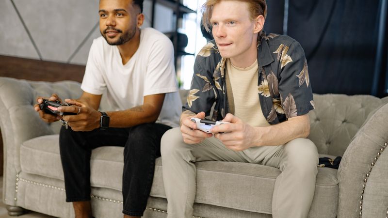 Two friends sitting on a sofa, playing video games together at home.