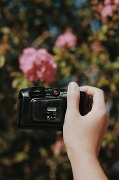 A close-up of a hand holding an analog camera against a blurred floral backdrop, highlighting natural photography themes.