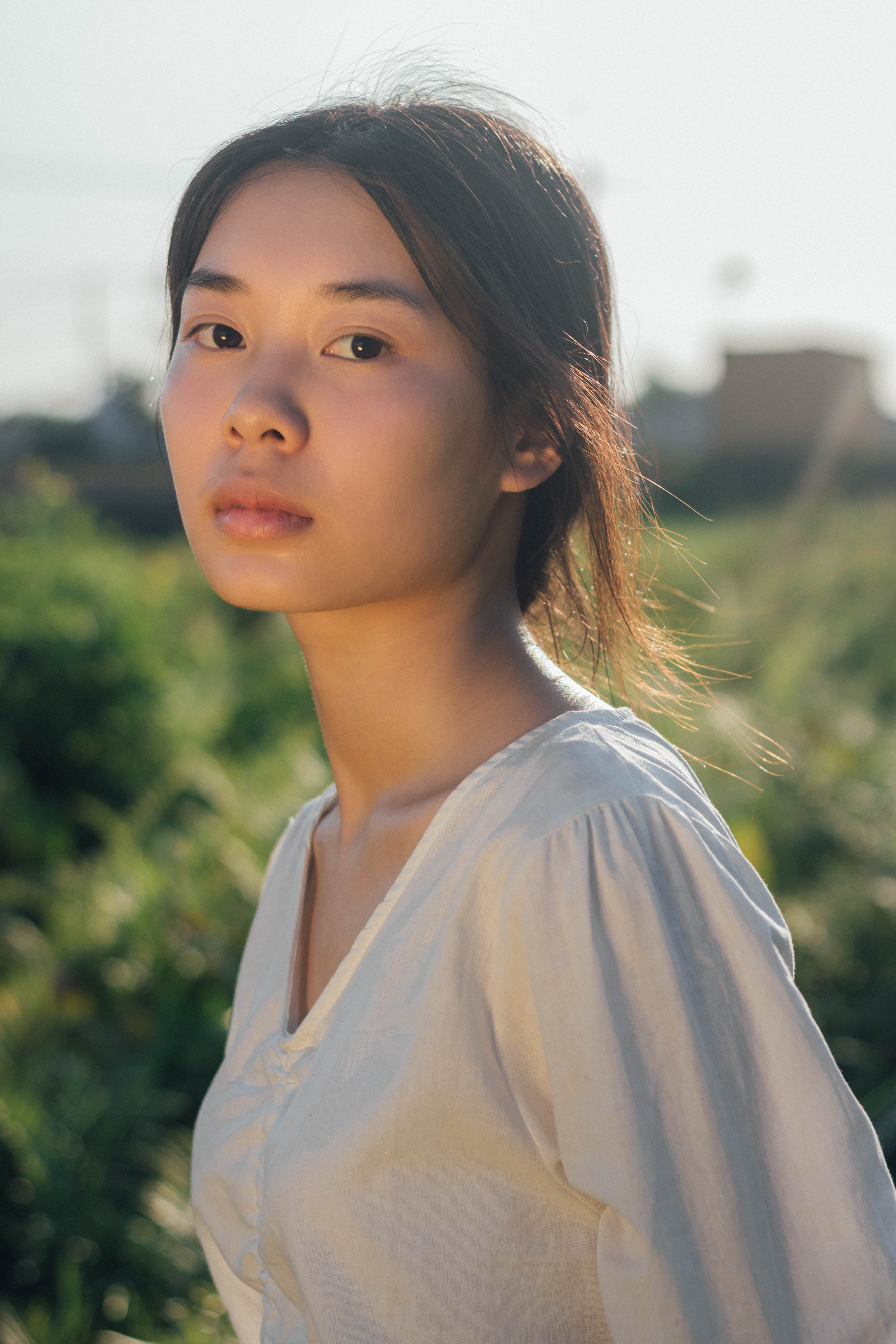 A thoughtful woman in a white blouse poses outdoors under natural daylight.