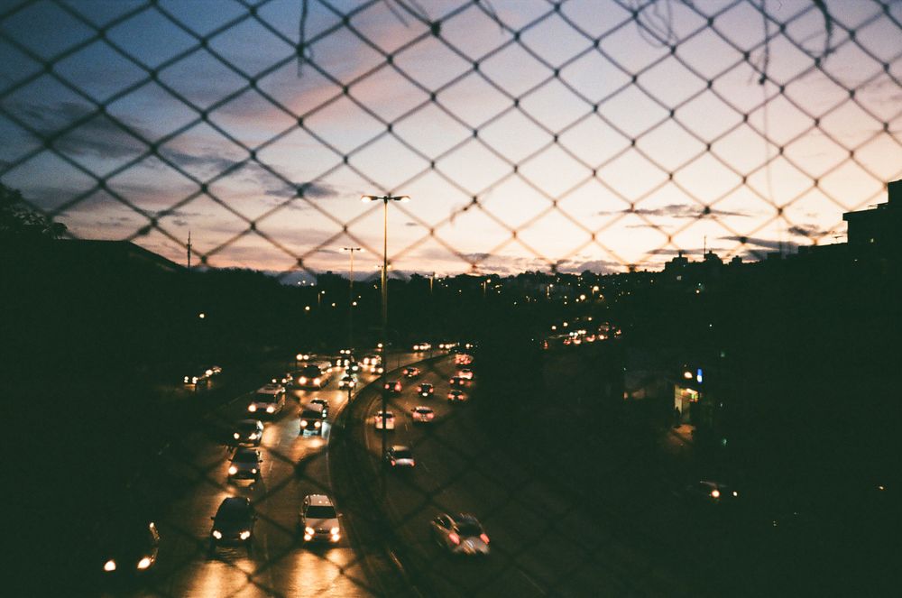 A view of city traffic captured through a chain link fence at dusk
