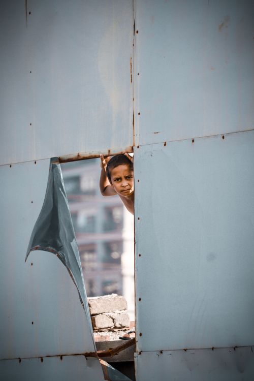 Free A curious child peeks through a torn metal sheet in an urban setting, showcasing innocence and curiosity. Stock Photo
