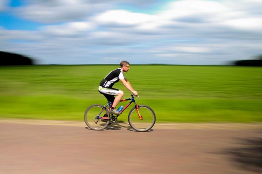 A cyclist riding at speed on a rural road with green fields and blue sky backdrop.