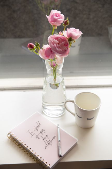 A glass vase with pink roses beside a notebook, pen, and cup on a windowsill.