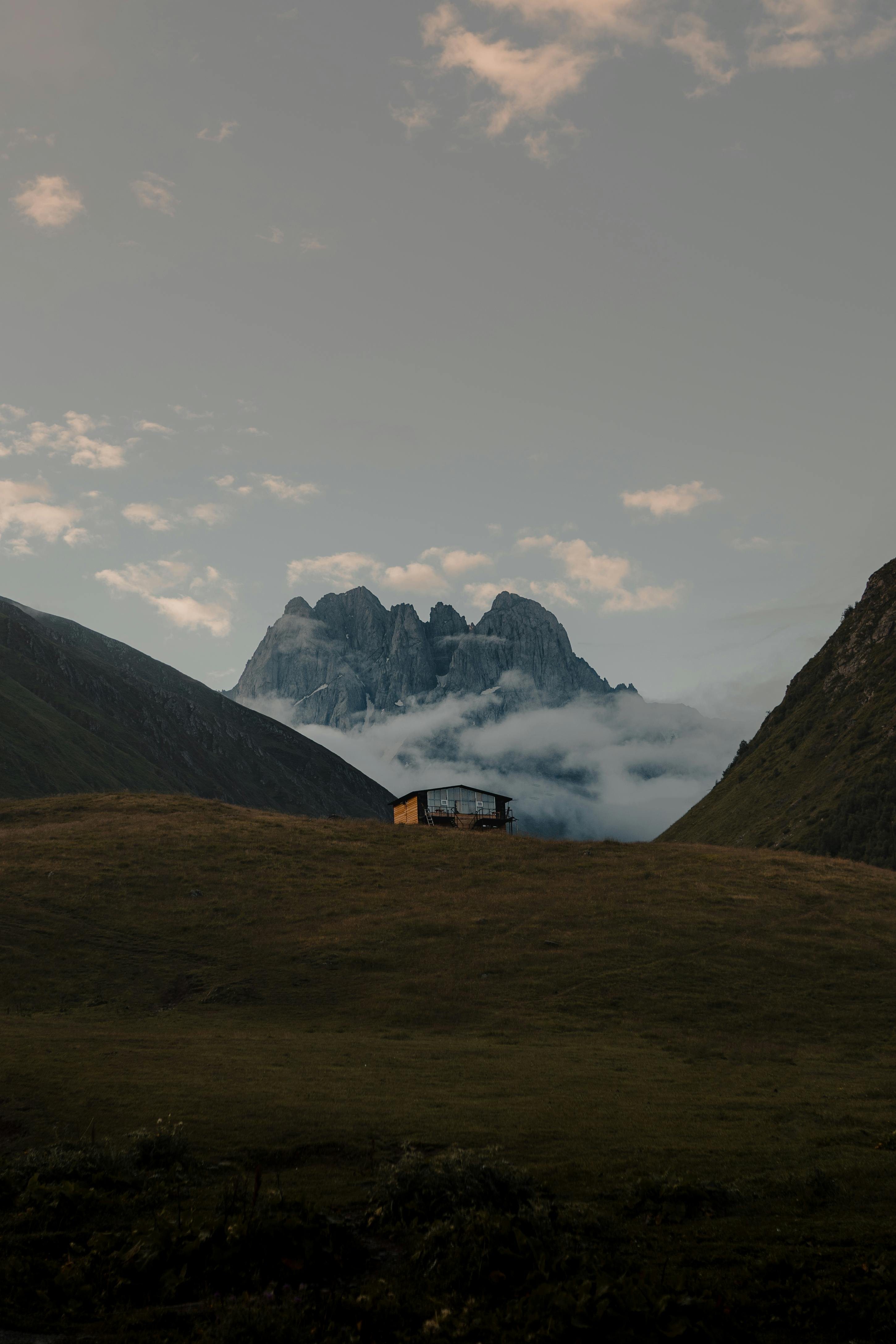 A cozy cabin on a hillside with towering mountains and misty clouds at dusk, offering a serene escape into nature.