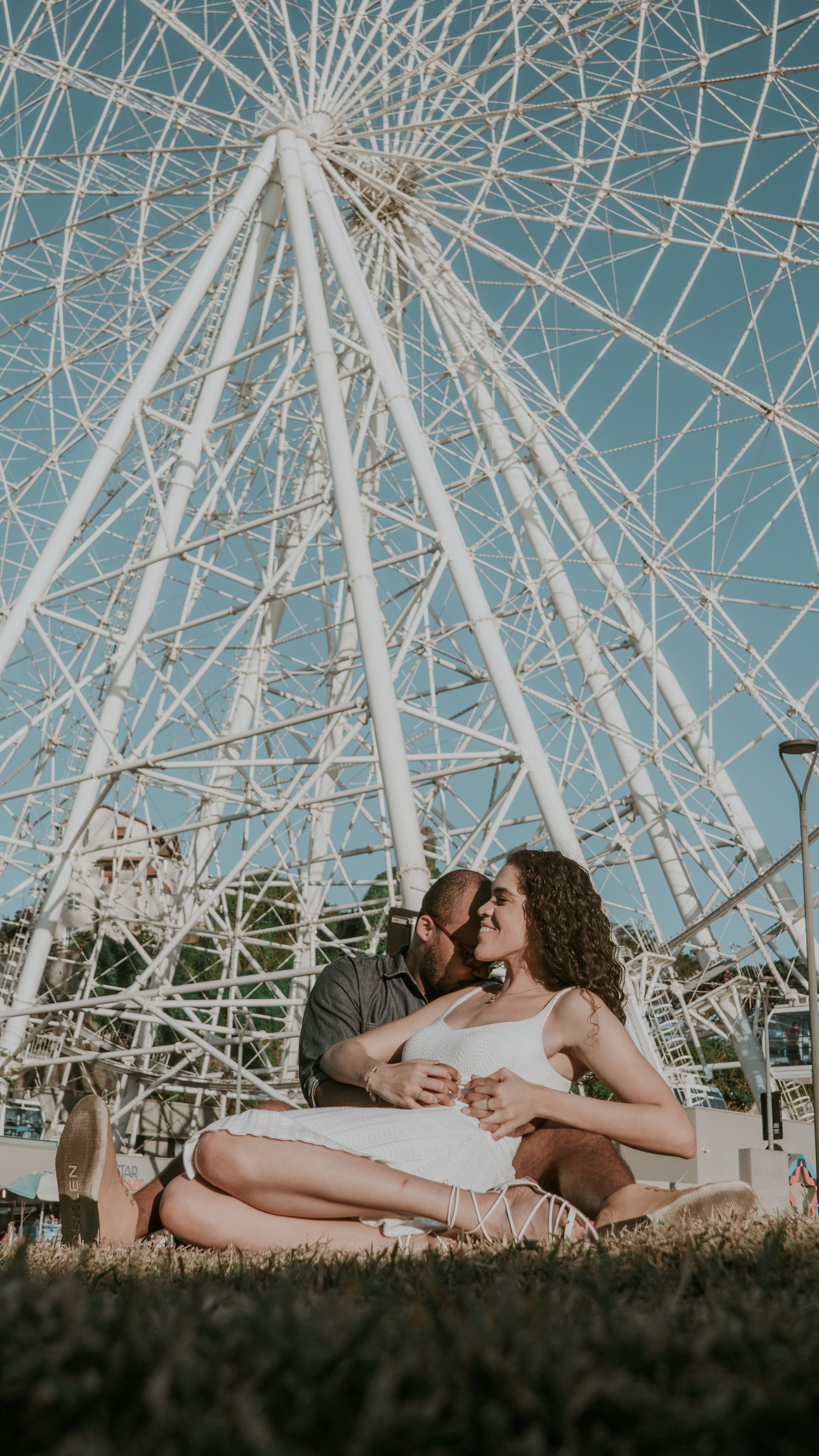 A romantic couple sharing a tender moment under a Ferris wheel during the day.