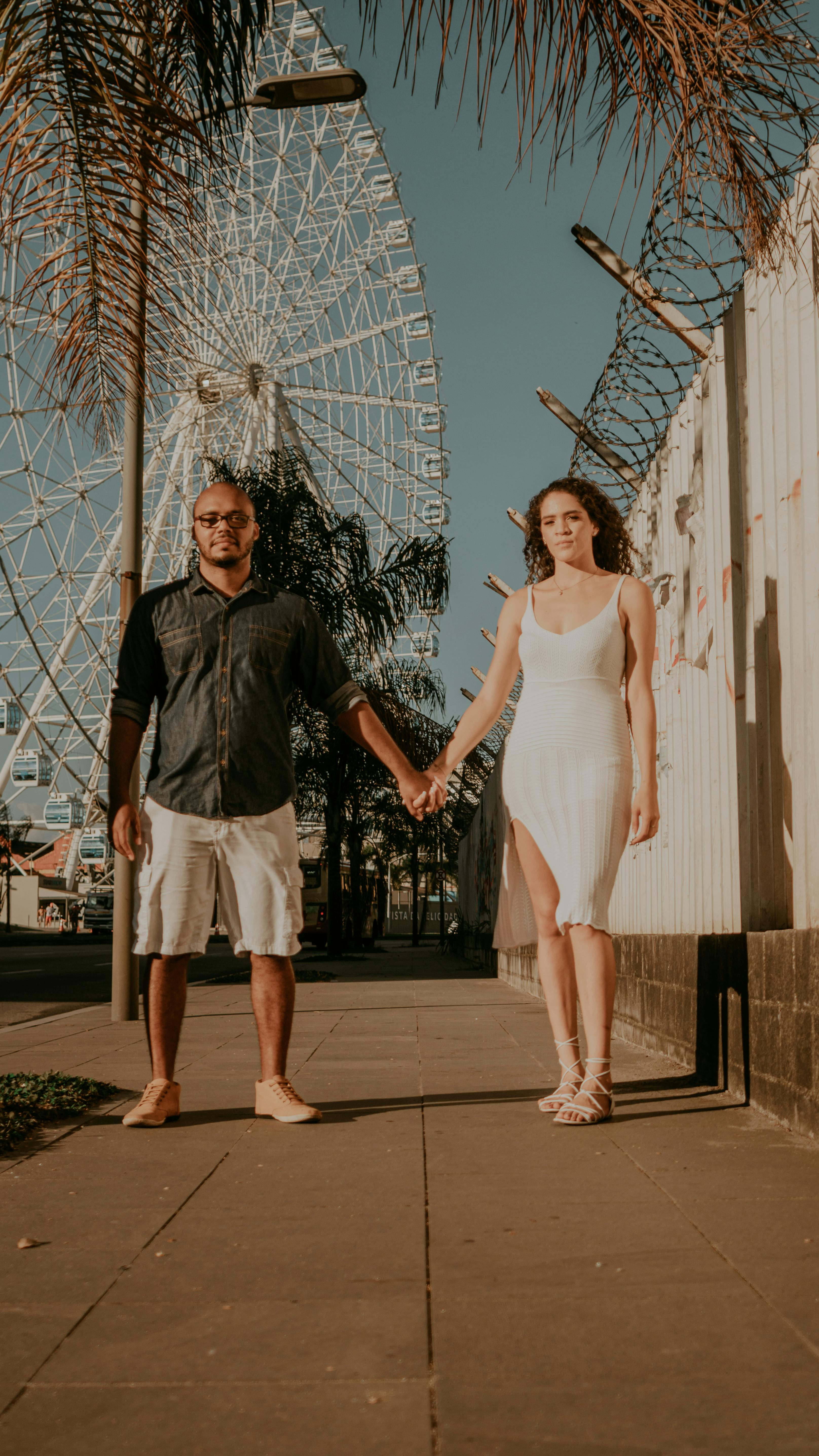 A couple holding hands on a sunny day near a Ferris wheel, symbolizing love and adventure.