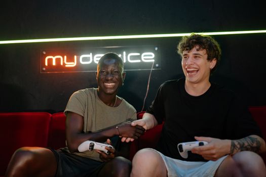 Two cheerful friends playing video games and shaking hands on a couch.