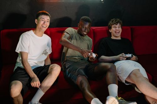 Free Three diverse young men sitting on a red couch, enjoying a fun video game session together. Stock Photo