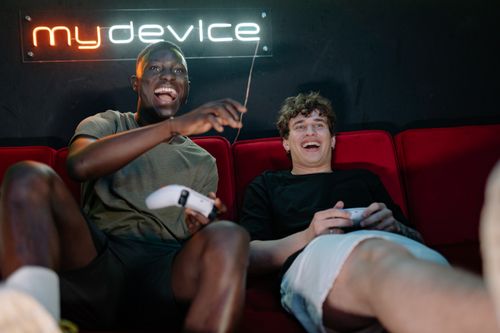 Free Two friends having fun playing video games, laughing and relaxing on a couch with a neon sign in the background. Stock Photo