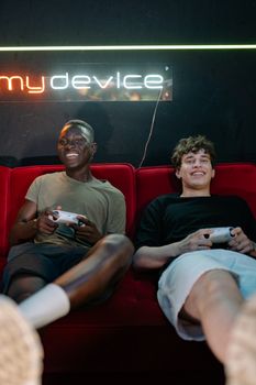 Two men playing video games with controllers on a red sofa, smiling indoors.