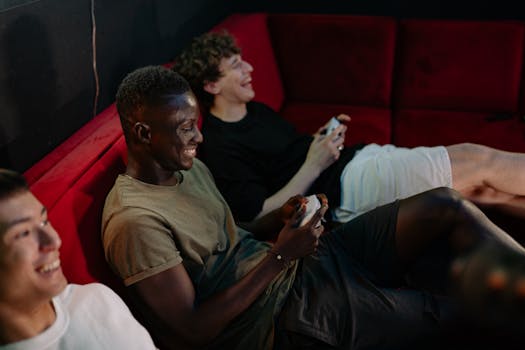 Three friends laughing and playing video games on a red sofa, having fun and relaxing indoors.
