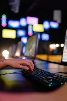 Hand typing on RGB gaming keyboard in a vibrant esports arena, showcasing colorful screens and gaming setup.