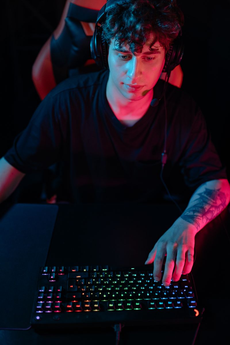A focused young man plays video games with RGB keyboard and headset at night.