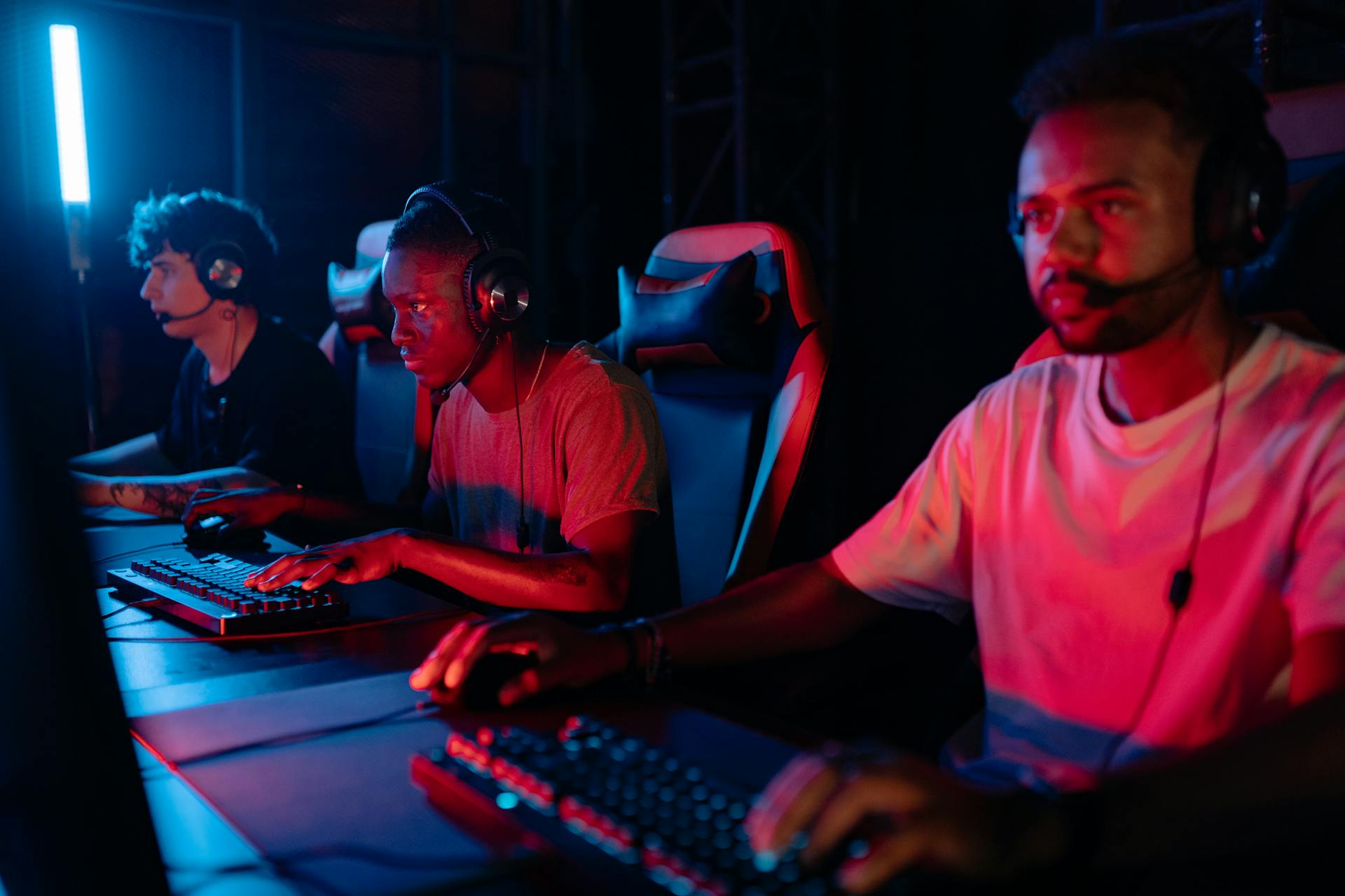 Three gamers immersed in a competitive setting with neon lighting, using keyboards and wearing headsets.