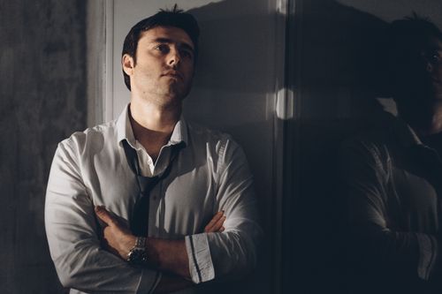 Free A thoughtful man in a white shirt leans against a wall, casting a shadow in dim light. Stock Photo