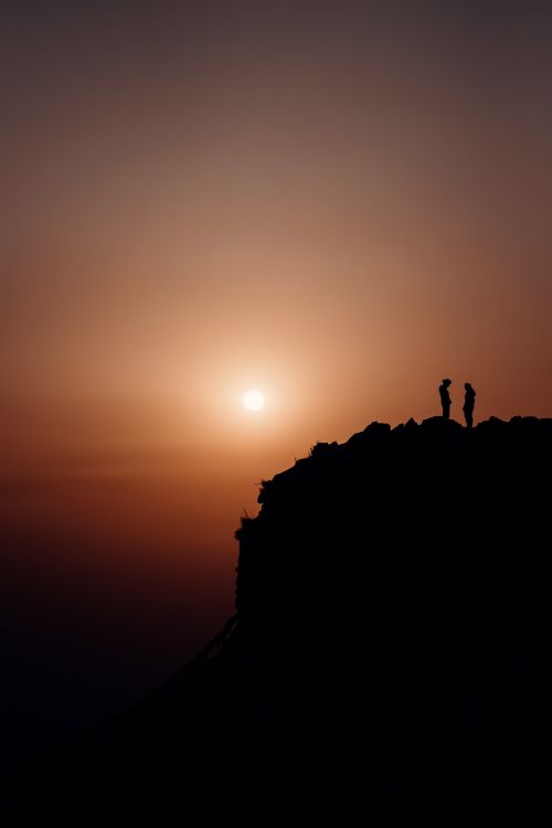 Free A silhouetted couple stands on a mountain ridge, basking in the golden sunrise over a vast landscape. Stock Photo