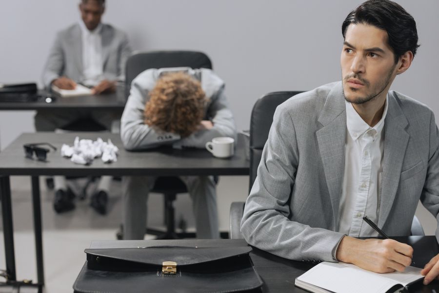 Office scene with employees in gray suits, one appearing bored and another taking notes.