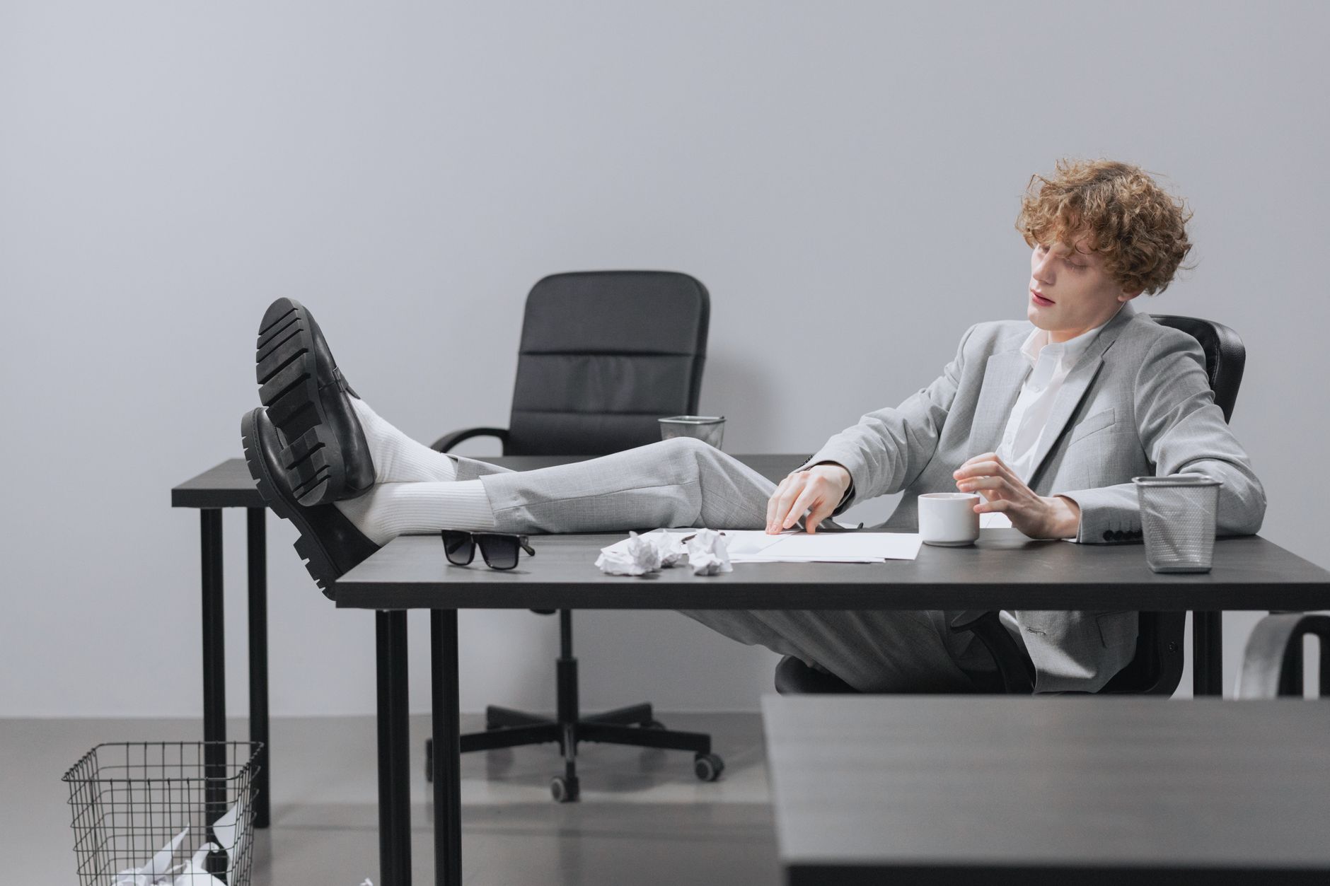 A curly-haired man in a suit relaxes with feet on desk, looking serious in a modern office setting.